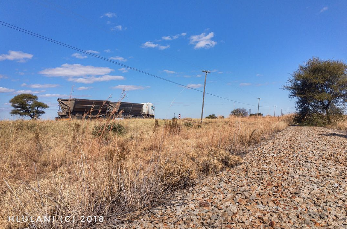 The photo shows a road freight truck along the vanishing point, with a stolen railway line on the lower end of the vanishing point. Between the two, there is a bush, trees and powerlines against the backdrop of bright blue Limpopo skies.